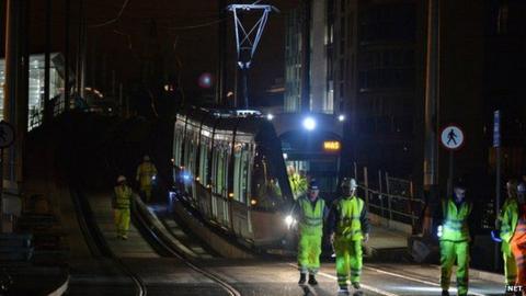 A tram crossing a bridge at night time with workers in high visibility clothing walking ahead of it.