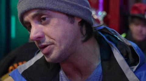 Jamie leans his head as he talks. He wears a grey beanie hat and blue coat over a grey top. He is in the room with other guests milling behind.