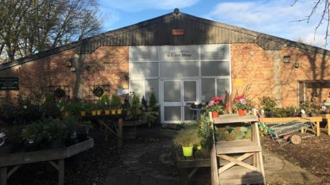 A large triangular brick building with a tinted glass door and windows that say "Darwin Nurseries & Farm Shop". In front of the building are several stalls with potted plants on them.