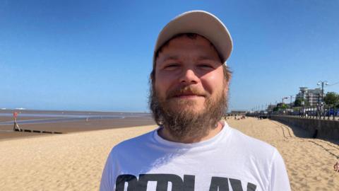 A head and shoulders view of a man stood on a beach. He is wearing a white cap and a white t-shirt with bold black writing on. He has a mid-length beard and moustache. Behind him is sand and blue skies.
