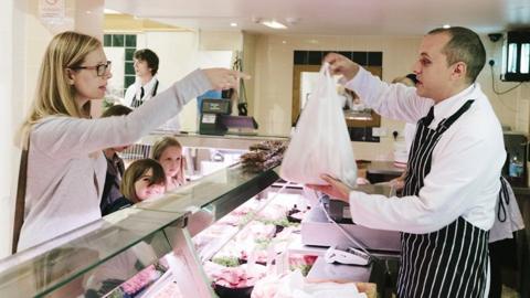 A file image of a woman at a butchers' counter with two small children, receiving a carrier bag of meat from a butcher wearing a striped apron 