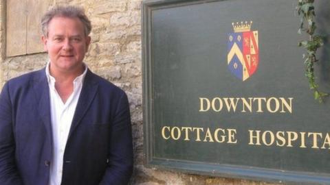 Hugh Bonneville next to a green sign reading 'Downton Cottage Hospital".