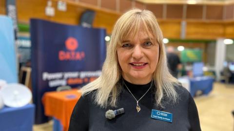 Helen Wylde-Chamberlain, CEO of Devon Chamber of Commerce, wearing a black sweater with a silver necklace. She has a small microphone on the left and a name tag on the right.
