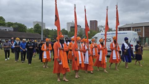 A group of people take part in a procession across a green lawn. The two front rows are made up of men wearing orange clothes and orange turbans. The five men at the very front are each holding an orange flag.