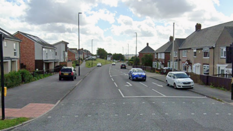 Old Mill Road is a three-lane road with two-storey houses lined on each side of it. There are a number of parked cars on the pavement and a pedestrian crossing. There are two cars travelling northbound.