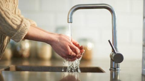 A silver tap running water with someone's hands cupped underneath it. They are wearing a woolly beige jumper and the background is blurred.