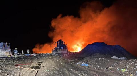 Firefighters and bulldozers at a large fire at a rubbish tip burning at night