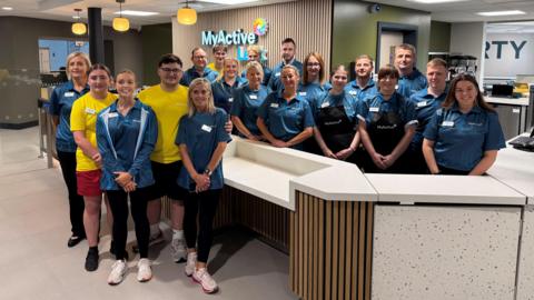 A group of people in blue or yellow uniforms are standing at a counter in a leisure centre with branding on the wall behind them. There is a pale grey flooring and a series of hanging lights dangling from the ceiling.