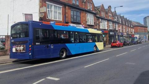 A blue and cream single-decker bus travels along a wide city street lined with red-brick terraced buildings featuring shopfronts and decorative rooflines. Several cars are parked, including a bright red car in the middle distance. On the left, a fenced car lot sits beside a wall. Modern high-rise apartment buildings are visible further down the road, contrasting with the older architecture. Overhead, the sky is mostly cloudy with patches of blue, and streetlights and road markings suggest a busy urban environment.
