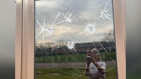 A picture of a smashed-up window, with the reflection of a woman taking a photograph of the damage.