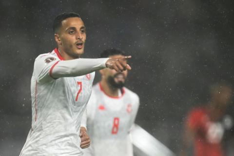 Footballer Elias Achouri celebrates after scoring a goal for Tunisia v Uganda at Afcon 2025. He is visible from chest up in a white away jersey and is extending his right arm straight out in front to point with his index finger. A team-mate is seen from neck up blurred in the background and large drops of rain can be seen falling