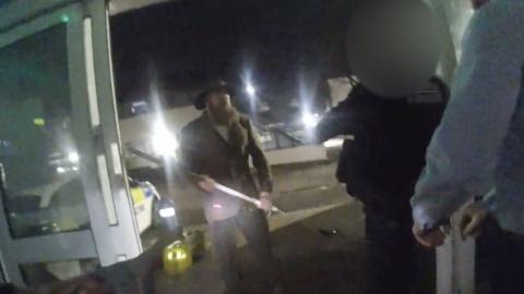 Man standing at the door to a police station, with a weapon in his hands.