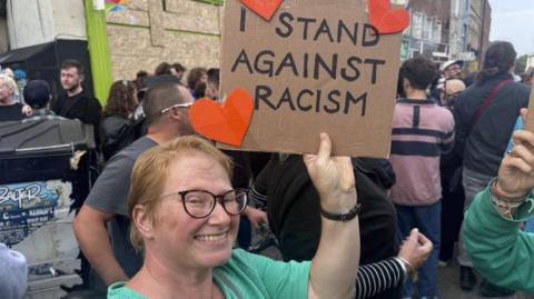 A lady in a green top at a rally in Bristol holding a sign that says 'I stand against racism'. There are also three red hearts on the sign. 