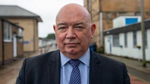 Michael Stoney in a blue short and suit with bald head looks straight at camera.