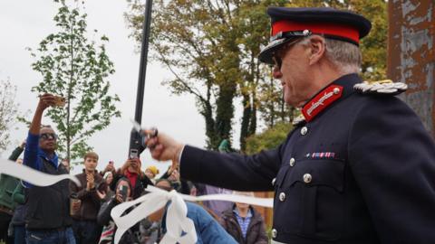 James Saunders Watson, the Lord Lieutenant of Northamptonshire, cutting a white ribbon, holding scissors in his hand, wearing a blue uniform, with a hat. He looking to the left. A crowd of people are in front of him, holding phones and clapping. Trees can also be seen.