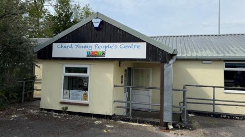 A one-storey, yellow building with a corrugated metal roof. There's a big sign  on front of the building. Metal railings surround the building, which has a car park in front of it.