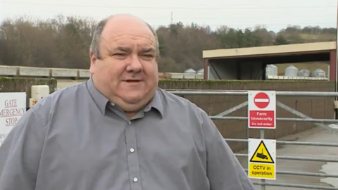 Mark Forster speaking to someone out of view in front of a farm gate. He has short grey hair thinning on the top of his head. He is wearing a grey shirt. 