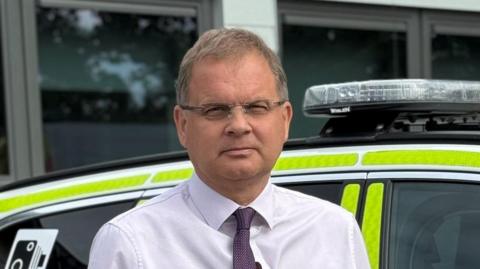Adam Pipe is wearing a white shirt, dark purple tie and glasses. He is standing in front of a police car.