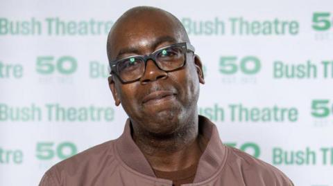 A black man with dark rimmed glasses smiles at the camera. He is wearing a brown coat at a film premiere. There is green writing on a white background behind him saying 'Bush Theatre 50'