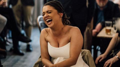 A woman wearing a white wedding dress is laughing while sat on a chair