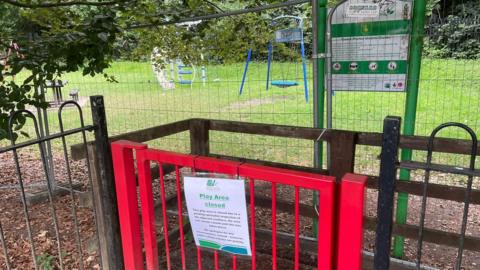 A gate to a playground with a notice attached, and temporary fencing between the entrance and the play equipment