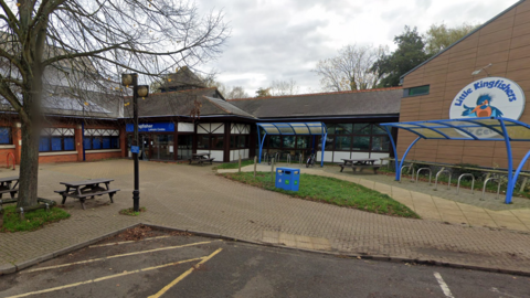 Bricked pool building to the left with a reception area in the centre of the photo. A large circular sign saying Little Kingfishers is on the right with an empty bike rack in front of it. There is also a litter bin and two picnic benches in the foreground.