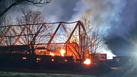 Outline of barn building on fire - the outline of the roof timbers can be seen and the sky is filled with large amounts of smoke.