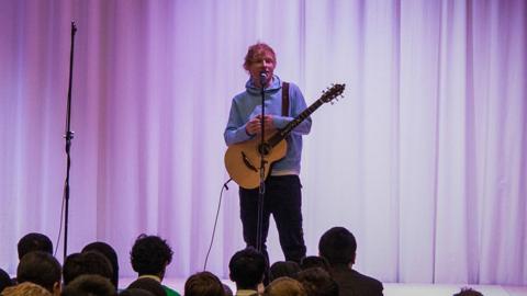 Ed Sheering on stage in front of school pupils. he is holding a guitar and is wearing a lights blue hoodie and dark trousers