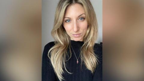 A woman with long hair and a dark coloured top. She is looking into the camera in the selfie.