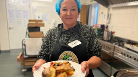 Volunteer cook Ann Wyatt holds a plate of vegetable stew and potato wedges as she stands in a kitchen, wearing an apron and a blue hairnet.