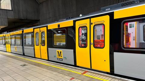 A yellow and grey Metro train pulled up at a platform. It has a sign in the window saying: "I'm being tested. Please do not attempt to board."