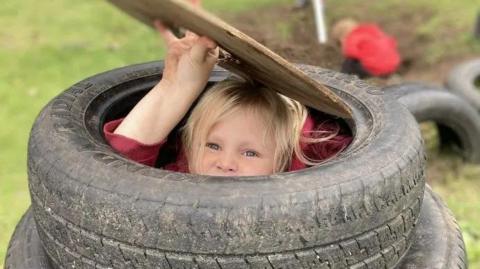  A child with blond hair is seen peeking out of play equipment made from  a mound of old car tyres   