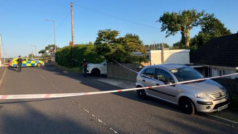 Police tape across a road in Ballymena. A silver car is parked at the roadside and a white vehicle is in a driveway. A police car is parked across the road at the rear of the picture and a police officer stands outside it