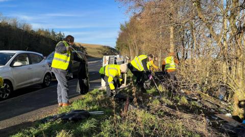 People in high visibility jackets gather litter from a banking with cars and a road in the background