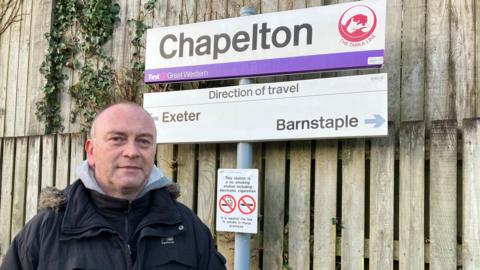 A  man with short hair and wearing a navy parker coat over a grey hoodie, He is standing in front of the Chapelton Train Station sign which is in front of a fence The signpost also includes a direction of travel sign pointing to Exeter on the left and Barnstaple on the right