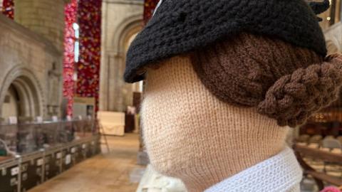 A close-up of the side view of a knitted female figure in World War Two dress in Peterborough Cathedral. It has a black hat, a cream face and brown hair in a bun. Beyond it are stone-flagged floors, cabinets with more knitted figures and stone arches.