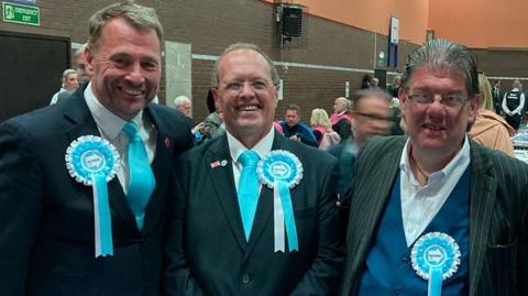 Stephen Atkinson, Ian Duxbury and Ged Mirfin at the Lancashire County Council count. All are wearing turquoise reform rosettes