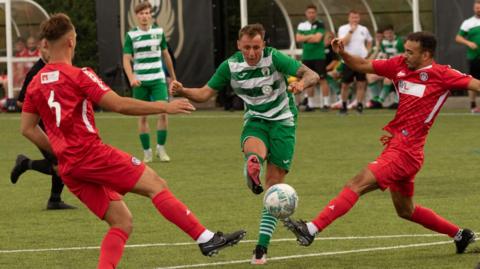 Three football players are going for the ball, two in red, one in a green and white shirt, while another player and the referee look on.