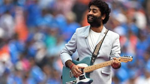 Arijit Singh performs in a grey suit jacket and white shirt while holding a blue guitar. He has a microphone attached to his head