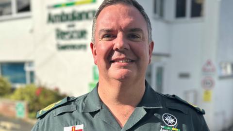 A man in a green ambulance service shirt smiles and looks towards us.