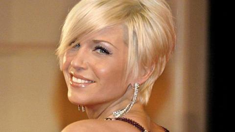 Sarah Harding, with blonde cropped hair and wearing diamond earrings and a purple strapped dress, looks over her shoulder and smiles