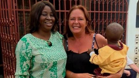 Two women standing outside and smiling at the camera. On the left is a teacher who has short brown hair and is wearing a green long-sleeved blouse. Next to her is Tracey Dennison who has long red hair. She is wearing a strappy black top and is holding a small child who is wearing a yellow top.