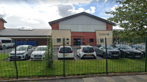 A street view of Woodland Community Primary School. It is a single story building with a car park, and a playground.