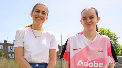 Two women on a football pitch. On the left, Karen Carney wears a white T-shirt. She has brown hair tied back and smiles. On the right, Ruby wears a pink branded sports T-shirt. She also has brown hair tied back. They are smiling.