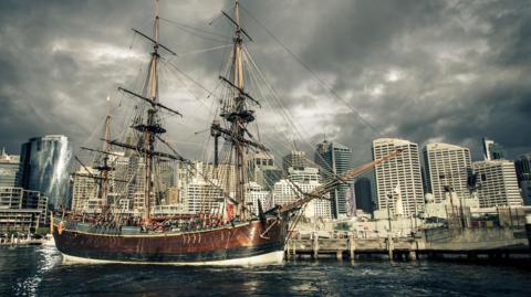 A three masted wooden sailing ship, about 100ft long, is docked in the harbour of a modern city, with tall office buildings in the background under a grey sky.