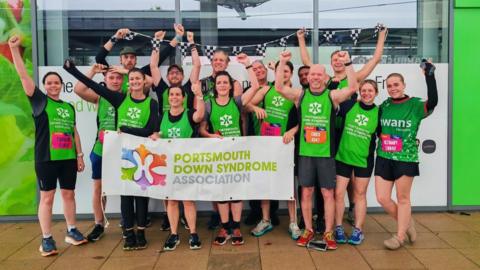 A group of runners with their hands in the air and they are wearing green vests.