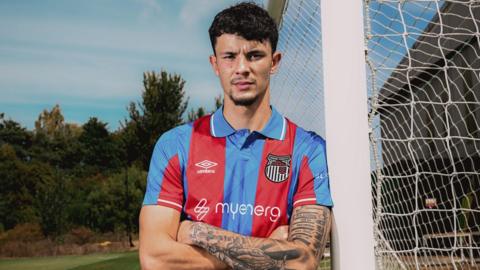 Jude Soonsup-Bell poses by leaning against a goalpost while wearing a Grimsby kit