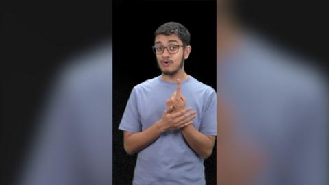 A man with a blue t shirt stood in front of a black background uses sign language.