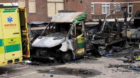 An ambulance that has been destroyed by fire. The front of the vehicle is intact but only a blackened skeleton remains of the remainder of it. Debris surrounds the vehicle.