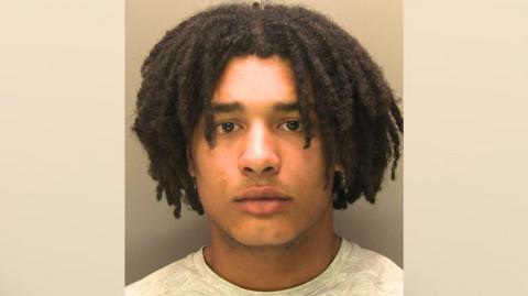 A police mugshot of a man with black hair wearing a grey t-shirt. He is in front of a blank wall looking at the camera. 